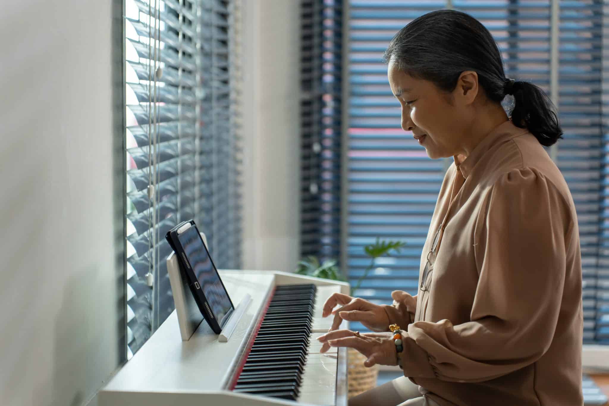 Asian senior mature woman play piano in living room at home. Beautiful elderly older grandmother feeling happy and relax while spend free leisure time learning music classroom from tablets.