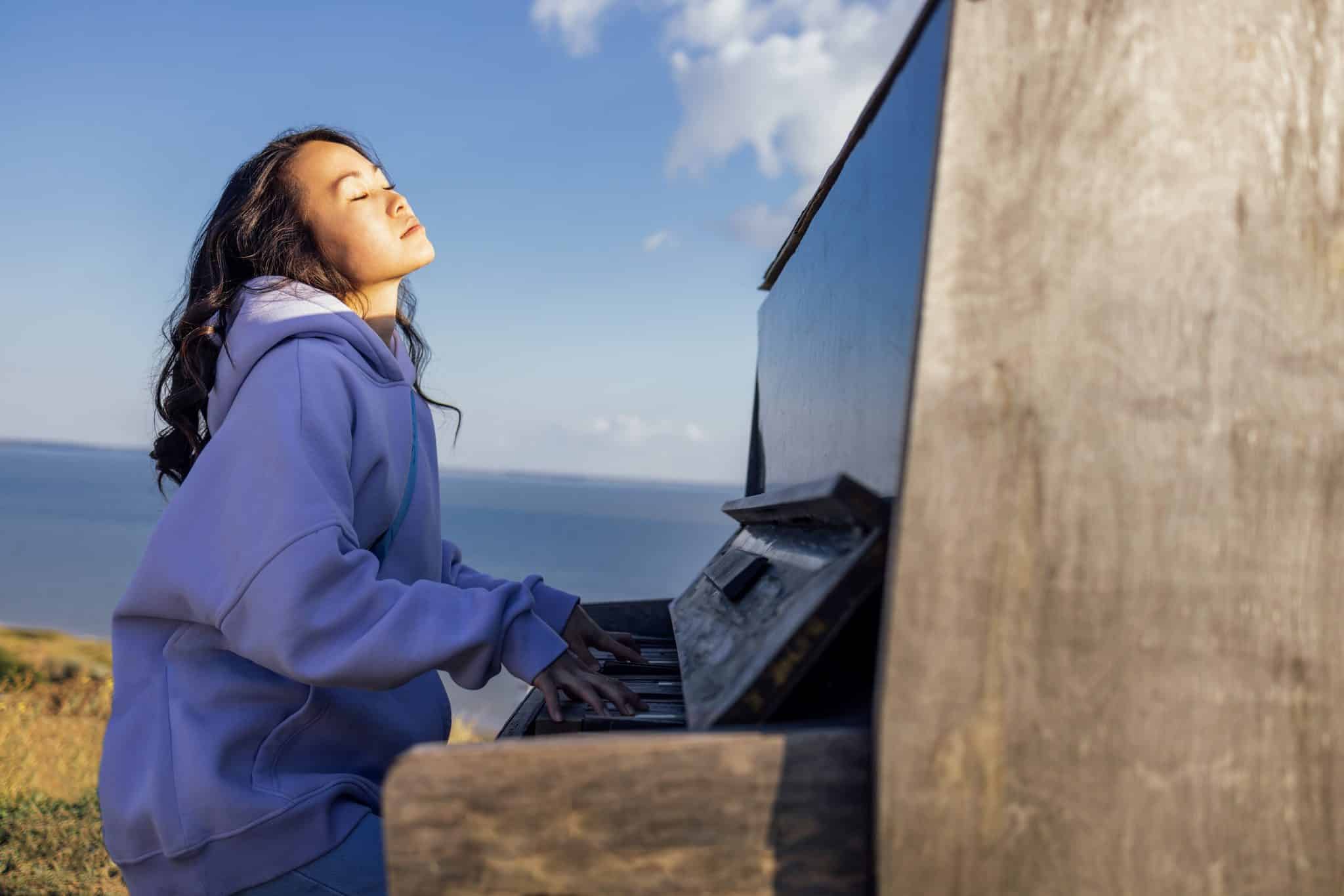 An Asian woman has closed her eyes and is playing the piano outdoors. A charming Korean girl enjoys playing a musical instrument in nature.