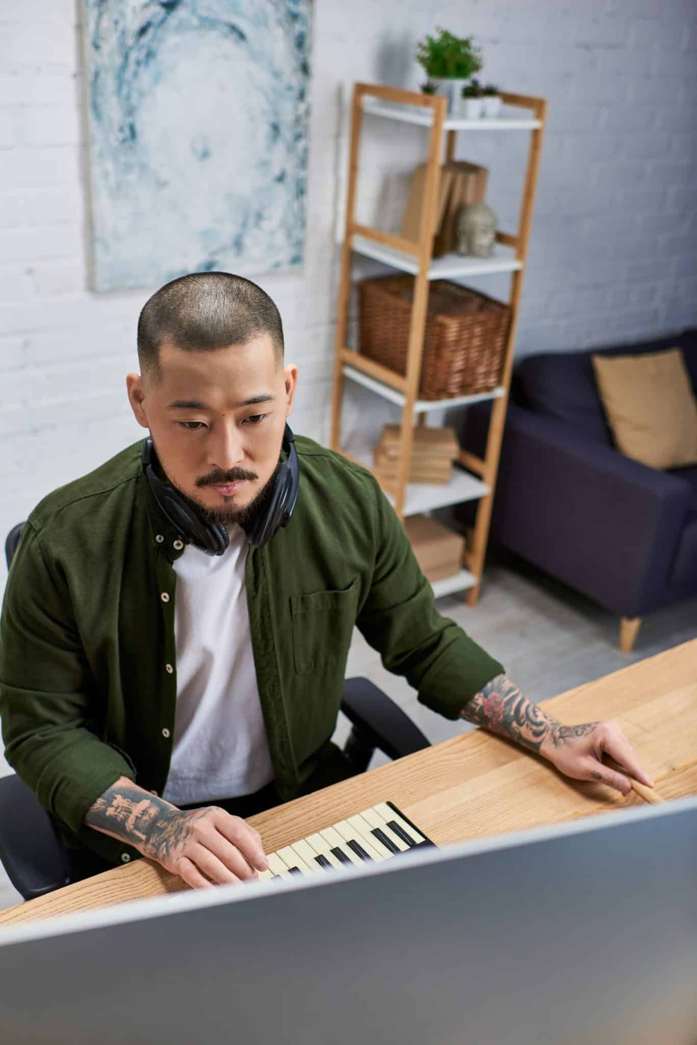 A handsome Asian man, wearing casual attire, sits at a desk in his studio, playing music on a keyboard and using a computer.