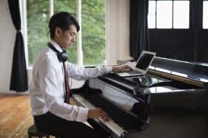 Young man sitting at a grand piano in a rehearsal studio, playing and simultaneously using a laptop computer.